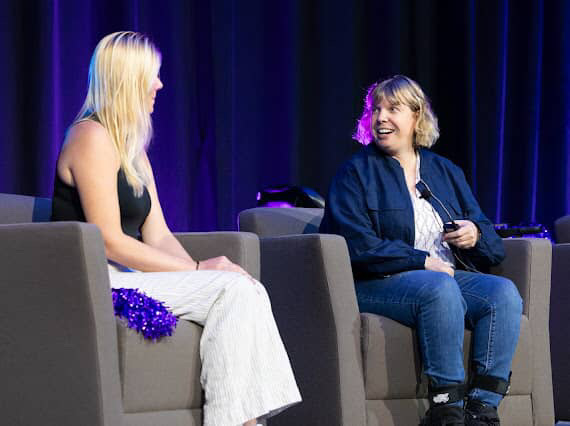 Darby smiling during a conversation with another speaker on a stage with blue curtains